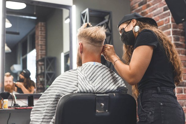 Female barber in mask cuts a man hairs with hair clipper. Hairstyle during social distancing. - Photo, Image