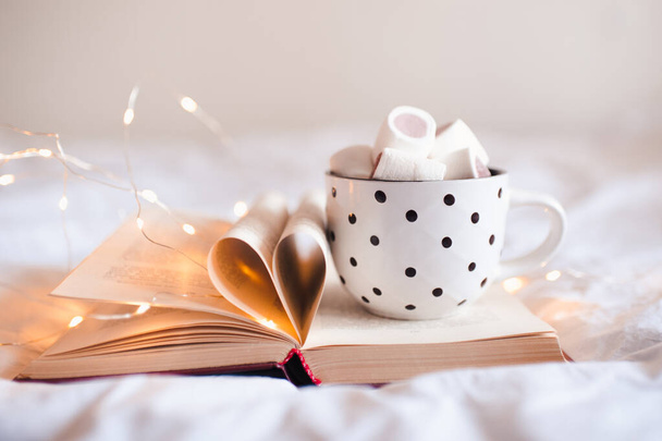Mug of coffee with marshmallow on open book and folded pages in heart shape closeup. Winter season.  - Photo, Image