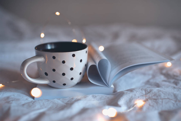 Tasse Tee auf aufgeschlagenem Buch mit herzförmigen gefalteten Seiten Nahaufnahme im Bett in der Nacht. Herbstzeit.  - Foto, Bild