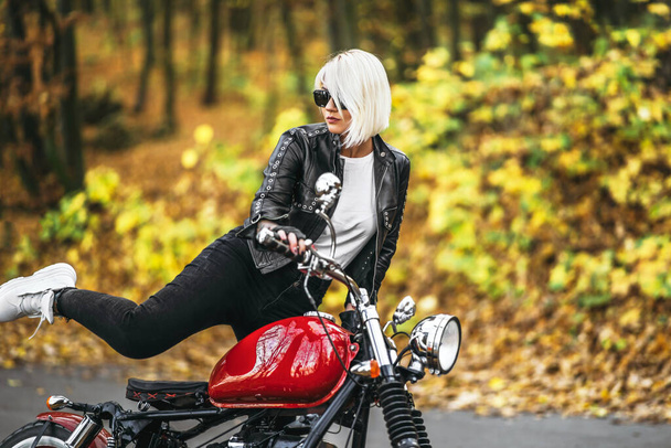 Pretty blonde biker girl in sunglasses with red motorcycle on the road in the forest with colorful blurred background. - Foto, Imagen