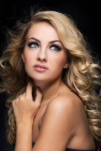 Beautiful woman with blonde curly hair posing with her shoulders naked over a black background - Фото, изображение