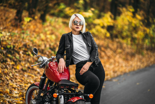 Pretty blonde biker girl in sunglasses with red motorcycle on the road in the forest with colorful blurred background. - Fotografie, Obrázek