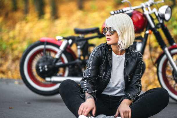 Pretty blonde biker girl in sunglasses sitting near red motorcycle on the road in the forest with colorful blurred background. - Foto, Imagen