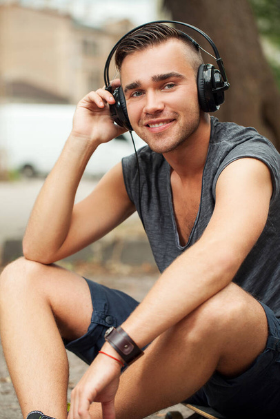 Portrait of a handsome man who is listening to music in his headphones while sitting in the street - Φωτογραφία, εικόνα