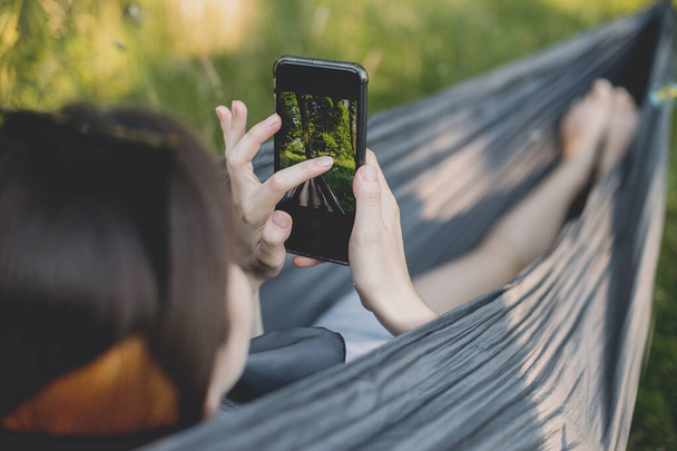 Young woman chilling in hammock and taking photo of her legs. Millennial girl holding phone and taking picture while relaxing in hammock, summer vacation - Photo, Image