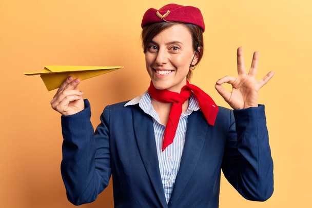 Jeune belle femme portant hôtesse uniforme tenant avion en papier faire ok signe avec les doigts, sourire geste amical excellent symbole  - Photo, image