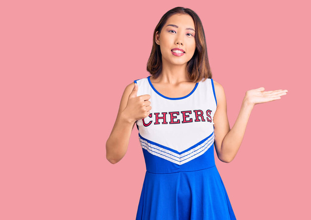 Joven hermosa chica china vistiendo uniforme de porrista mostrando la palma de la mano y haciendo buen gesto con los pulgares hacia arriba, sonriendo feliz y alegre  - Foto, Imagen
