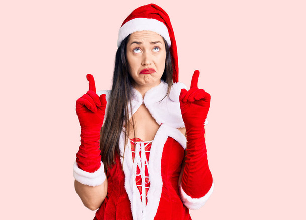 Junge schöne kaukasische Frau im Weihnachtsmann-Kostüm, die traurig und aufgebracht nach oben zeigt, mit den Fingern die Richtung anzeigt, unglücklich und deprimiert.  - Foto, Bild