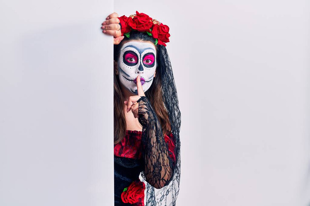 Jonge vrouw draagt dag van de dode custome houdt lege banner en vraagt om stil te zijn met de vinger op de lippen. stilte en geheim concept.  - Foto, afbeelding