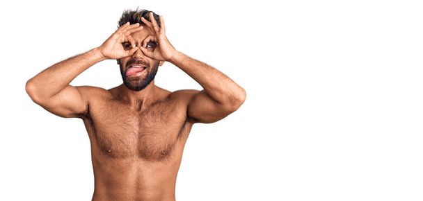 Un joven hispano con traje de baño sin camisa haciendo un buen gesto como binoculares sacando la lengua, ojos mirando a través de los dedos. expresión loca.  - Foto, Imagen