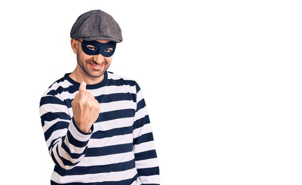 Junger gutaussehender Mann mit Einbrechermaske winkt hier mit Handzeichen freundlich lächelnd ein  - Foto, Bild