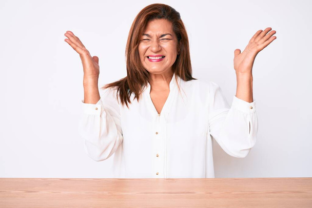 Middelbare leeftijd brunette Latijns-Amerikaanse zakenvrouw dragen casual wit shirt zitten op de tafel vieren gek en gek op succes met armen omhoog en gesloten ogen schreeuwend opgewonden. winnaarsconcept  - Foto, afbeelding