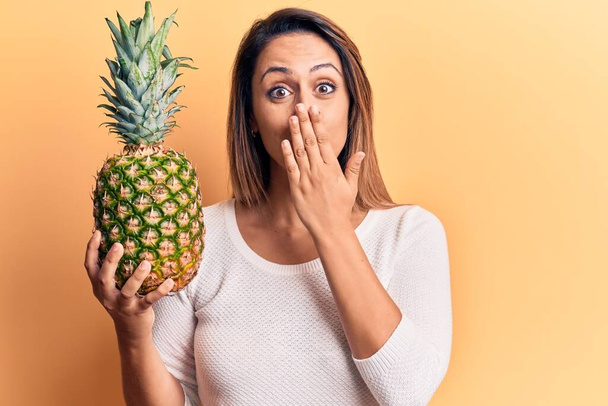 Nuori kaunis nainen pitelee ananas peittää suun käsin, järkyttynyt ja pelkää erehdystä. yllättynyt ilme  - Valokuva, kuva