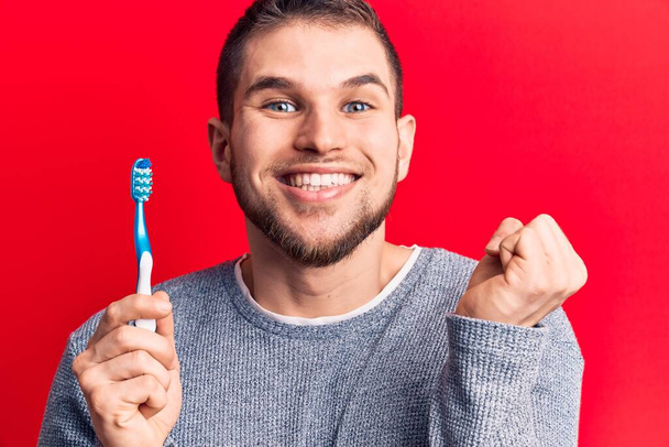 Young handsome man holding toothbrush screaming proud, celebrating victory and success very excited with raised arm  - Zdjęcie, obraz