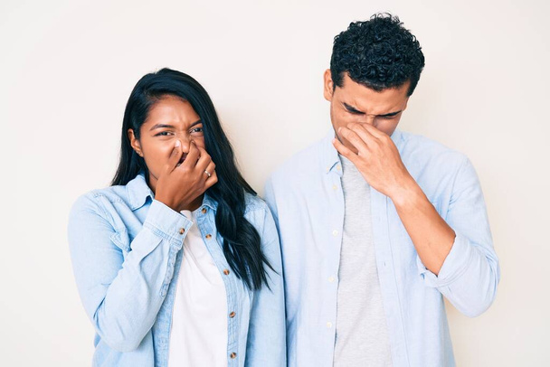 Beautiful latin young couple wearing casual clothes smelling something stinky and disgusting, intolerable smell, holding breath with fingers on nose. bad smell  - Foto, Imagen