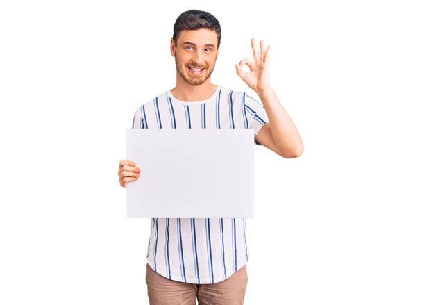 Jóképű fiatalember medvével a kezében üres banner csinál rendben jel ujjakkal, mosolygós barátságos gesztus kiváló szimbólum  - Fotó, kép