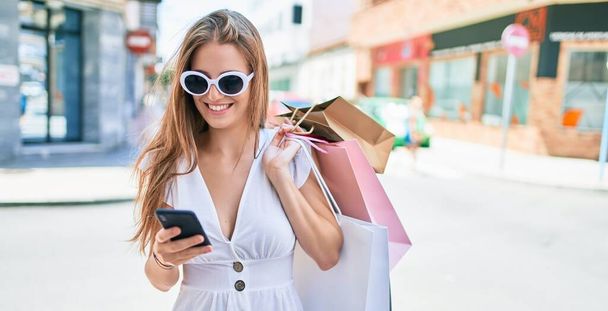 Mujer rubia joven sonriendo feliz sosteniendo bolsas de compras y el uso de teléfono inteligente en la calle de la ciudad - Foto, Imagen