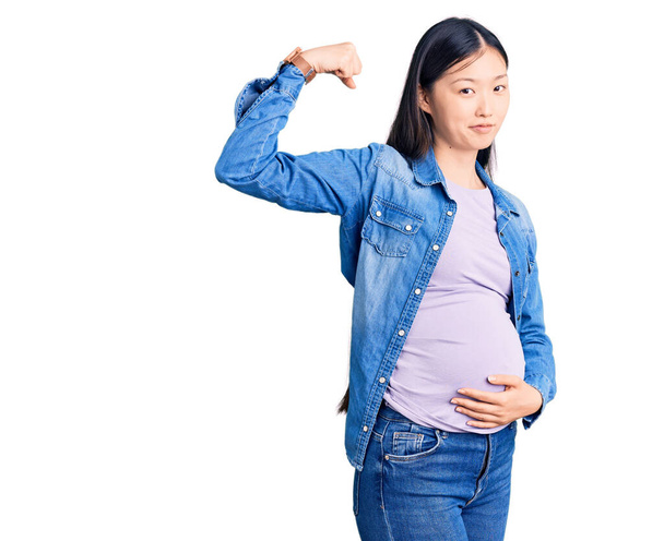 Junge schöne chinesische Frau schwanger erwartet Baby starke Person zeigt Armmuskeln, selbstbewusst und stolz auf Macht  - Foto, Bild