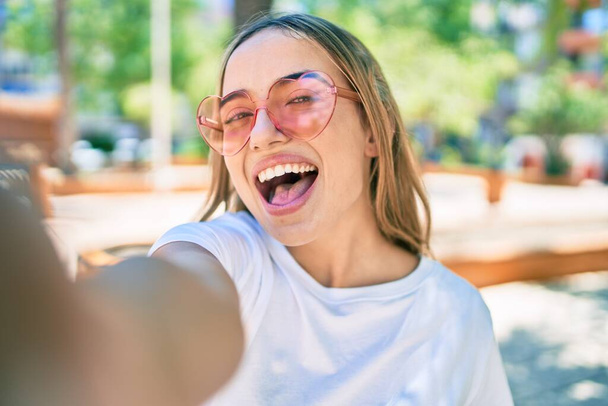 Jeune belle blonde caucasienne souriante heureuse en plein air par une journée ensoleillée portant des lunettes de soleil en forme de coeur prenant une photo selfie - Photo, image