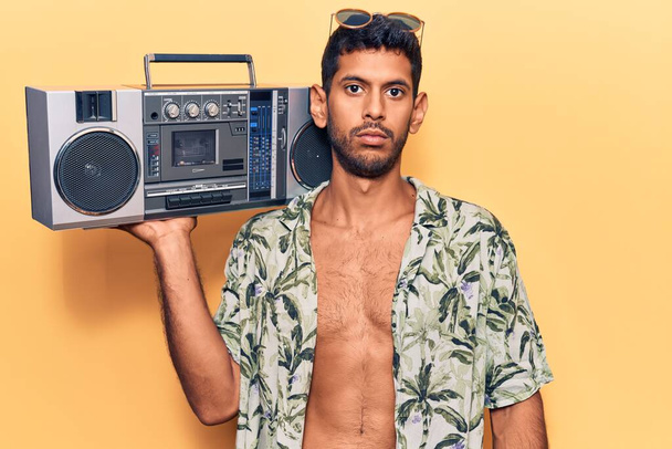 Mladý latinský muž v letní košili drží boombox myšlení postoj a střízlivý výraz hledá sebevědomí  - Fotografie, Obrázek