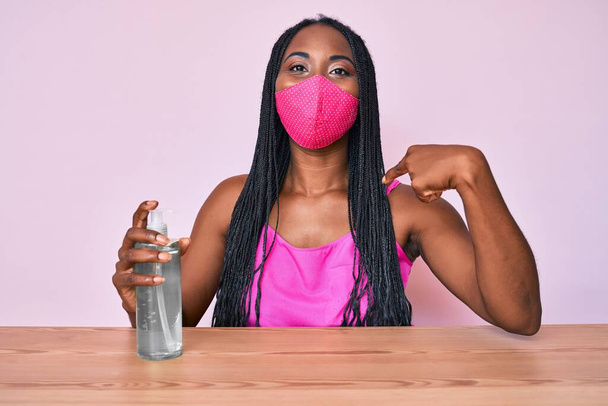 African american woman with braids wearing medical mask holding hand sanitizer gel pointing finger to one self smiling happy and proud  - Photo, Image