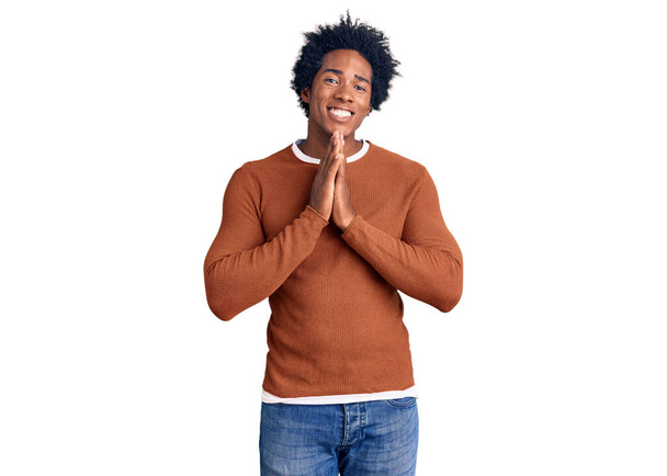 Красивый африканский американец с афроволосами в повседневной одежде молится руками, прося прощения, уверенно улыбаясь.  - Фото, изображение