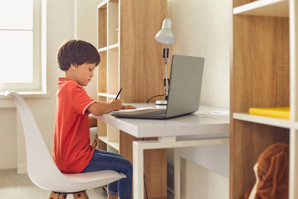 Маленький хлопчик використовує ноутбук, щоб зробити домашнє завдання, сидячи за столом у своїй кімнаті
. - Фото, зображення