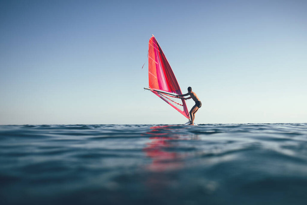 Nuori mies nostaa purjeita purjelautailuun auringonlaskun merelle. Siluetti purjelautailija balacning surffilauta - Valokuva, kuva