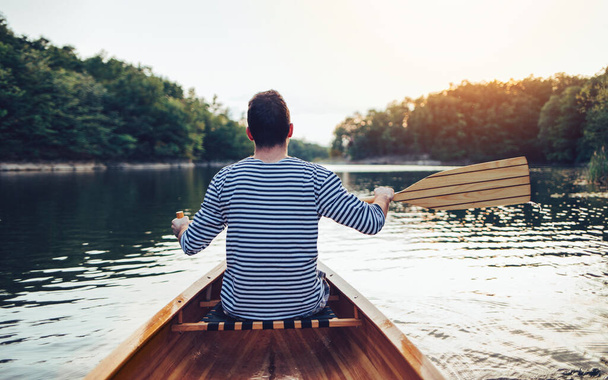 Man in sailor shirt paddling boat on the sunset lake. Canoeing, paddling, water sport - Foto, Bild