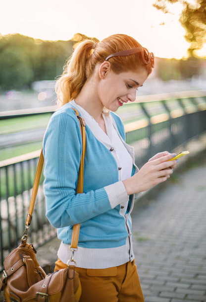 Młoda kobieta pisze SMS-y na smartfonie. Uśmiechnięta kobieta smsuje smsy w parku - Zdjęcie, obraz