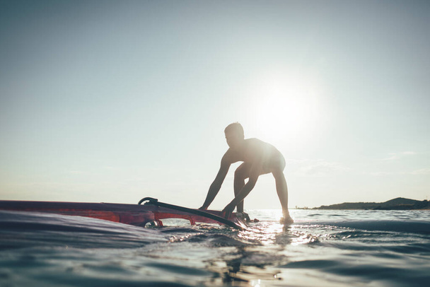Young man uplift windsurf board sail. Surfer balancing on wind surf board on sunset sea. Windsurfing, summer, surfing, lifestyle - Photo, Image