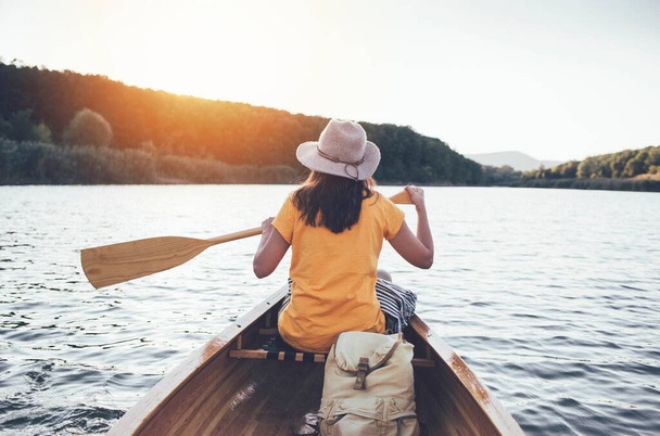 Rear view of girl with hat paddling canoe on sunset lake - Фото, зображення