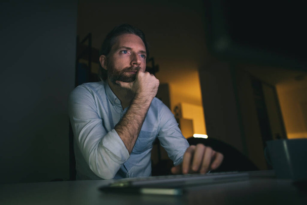 Hombre barbudo trabajando hasta tarde en la computadora. Hombre trabajando largas horas desde su oficina en casa - Foto, Imagen