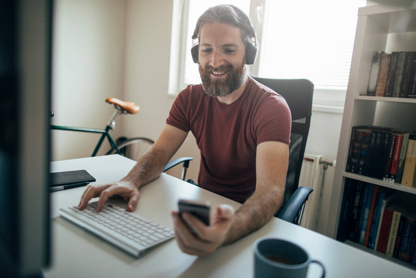 Sonriente hombre barbudo sosteniendo teléfono inteligente en su oficina en casa. Hombre trabajando desde casa - Foto, imagen