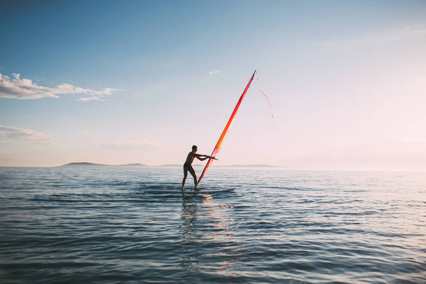 Mladý muž zvedá plachtu na windsurf. Surfař balancující na windsurf palubě na západě slunce moře. Windsurfing, léto, surfování, životní styl - Fotografie, Obrázek