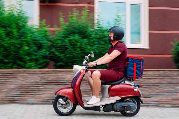 Delivery boy wearing red uniform on scooter with isothermal food case box driving fast. Express food delivery service from cafes and restaurants. Courier on the moto scooter delivering food. - 写真・画像