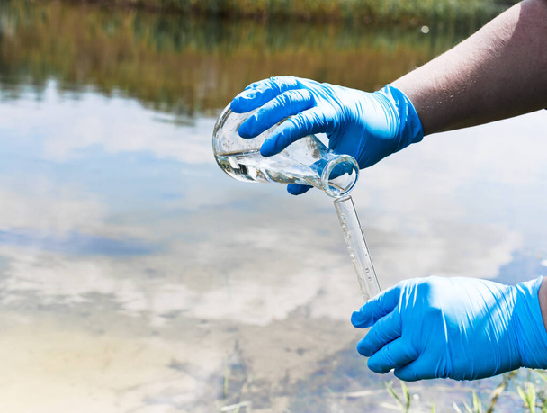 Valla, revisa la muestra de agua en busca de infecciones. Guante de mano con un matraz y un tubo de ensayo sobre un fondo de lago, río, mar. Contaminación de la ecología, medio ambiente - Foto, Imagen
