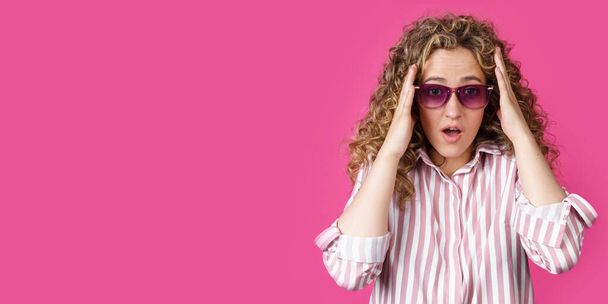 A young woman holds her head with her hands, surprised. Isolated on pink background - Фото, изображение