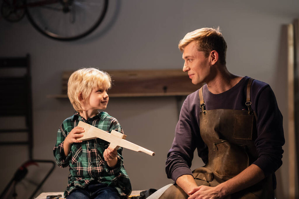 een jonge timmerman vader en zijn schattige blonde zoon maakte een pistool van hout en tonen het, ze zijn gelukkig, zitten in de timmermanswerkplaats op de tafel. - Foto, afbeelding