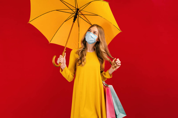 Pensive beautiful young woman in a medical protective mask, with shopping bags and umbrella, on a red background, shopping concept - Photo, image