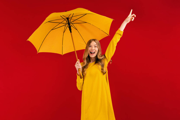 Excited happy young woman with a yellow umbrella having fun and showing a winning gesture on a red background, autumn concept - Фото, изображение