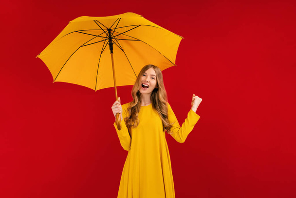 Excited young woman with a yellow umbrella having fun and giving a winning gesture on a red background, autumn concept - Фото, изображение