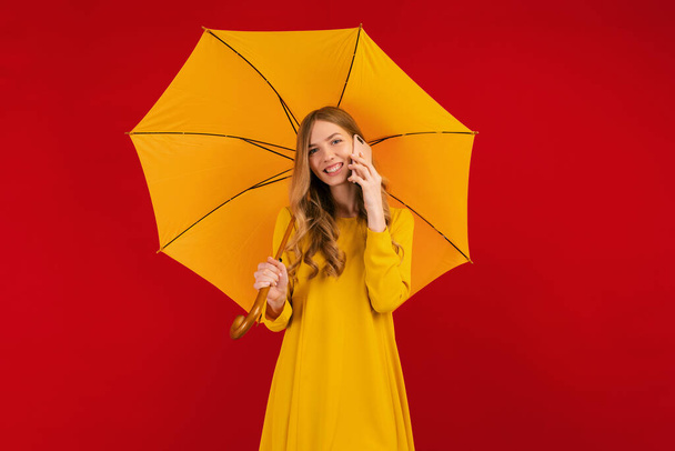 Piękna szczęśliwa młoda kobieta w żółtej sukience z parasolem, rozmawiająca przez telefon komórkowy na czerwonym tle, koncepcja jesienna - Zdjęcie, obraz