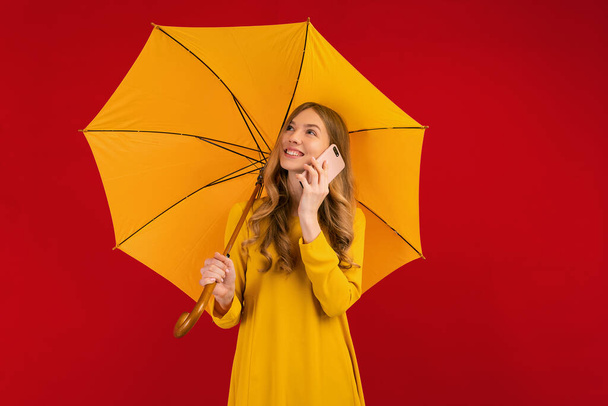 Beautiful happy young woman in a yellow dress with an umbrella, talking on a mobile phone on a red background, autumn concept - Foto, Imagen