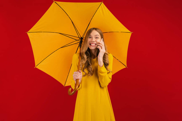 Beautiful happy young woman in a yellow dress with an umbrella, talking on a mobile phone on a red background, autumn concept - Photo, Image