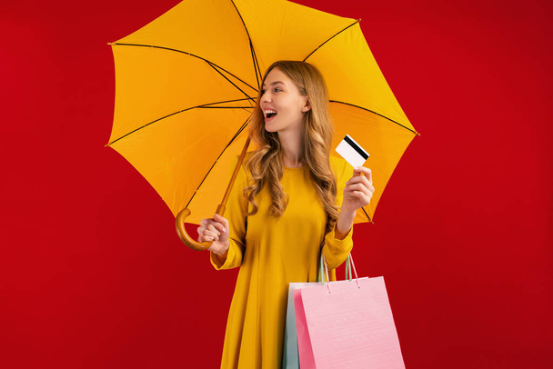 Happy beautiful young woman with umbrella holding shopping bags and credit card on red background - Zdjęcie, obraz