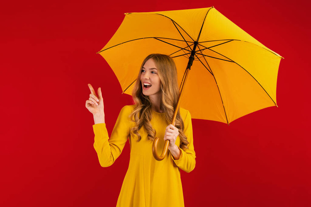 happy young woman with a yellow umbrella pointing at a copy of the space on an empty red background - Valokuva, kuva
