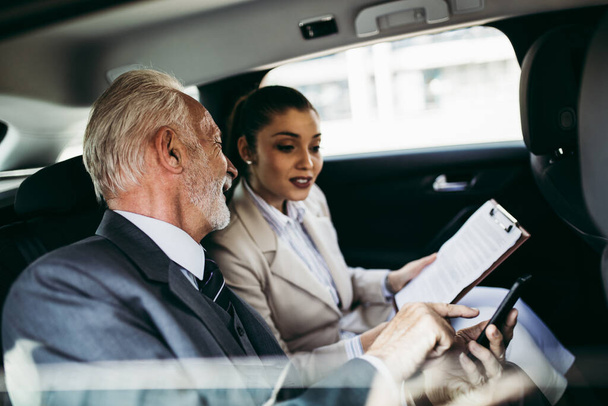 Good looking senior business man and his young woman colleague or coworker sitting on backseat in luxury car. They talking, smiling and using laptop and smart phones. Transportation in corporate business concept. - Photo, image