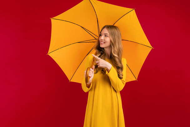 happy young woman with a yellow umbrella pointing at a copy of the space on an empty red background, autumn concept - Фото, изображение