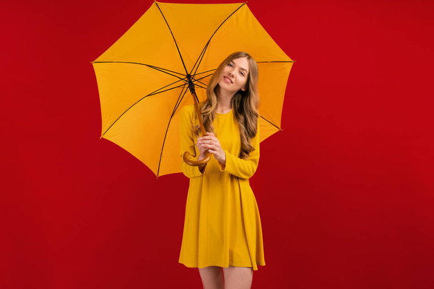 Beautiful happy young woman with yellow umbrella on red background, autumn concept - Fotó, kép